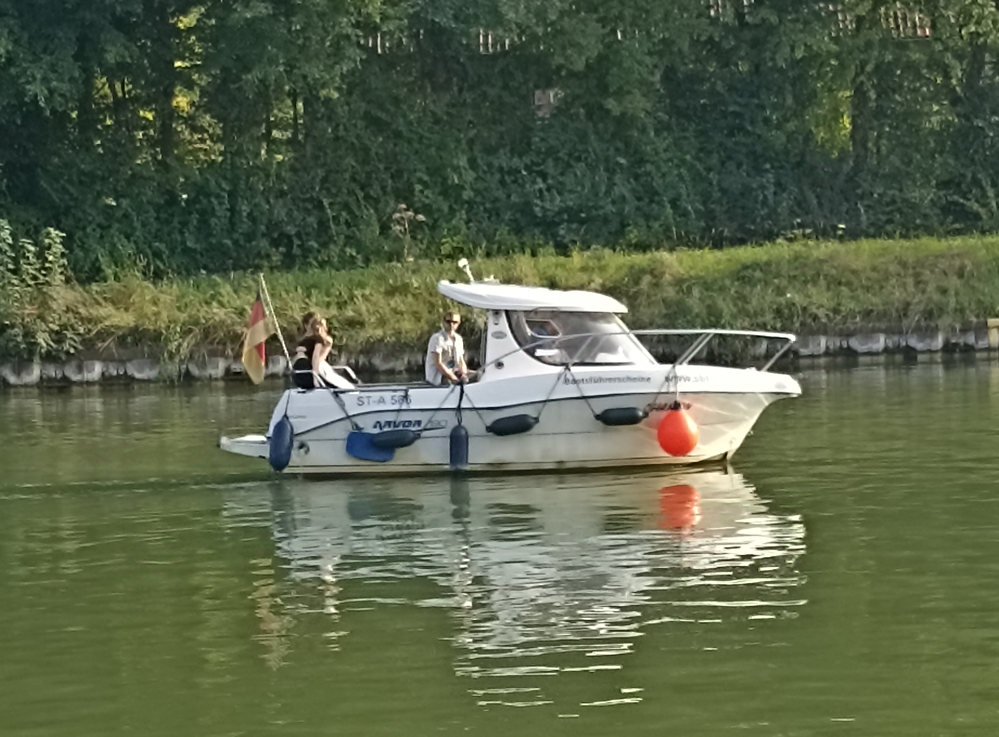 Ein weißes Sportboot mit Fendern fährt nach rechts, besetzt mit drei entspannt wirkenden Personen, im Hintergrund sind Bäume eine Spundwand ist zu erkennen