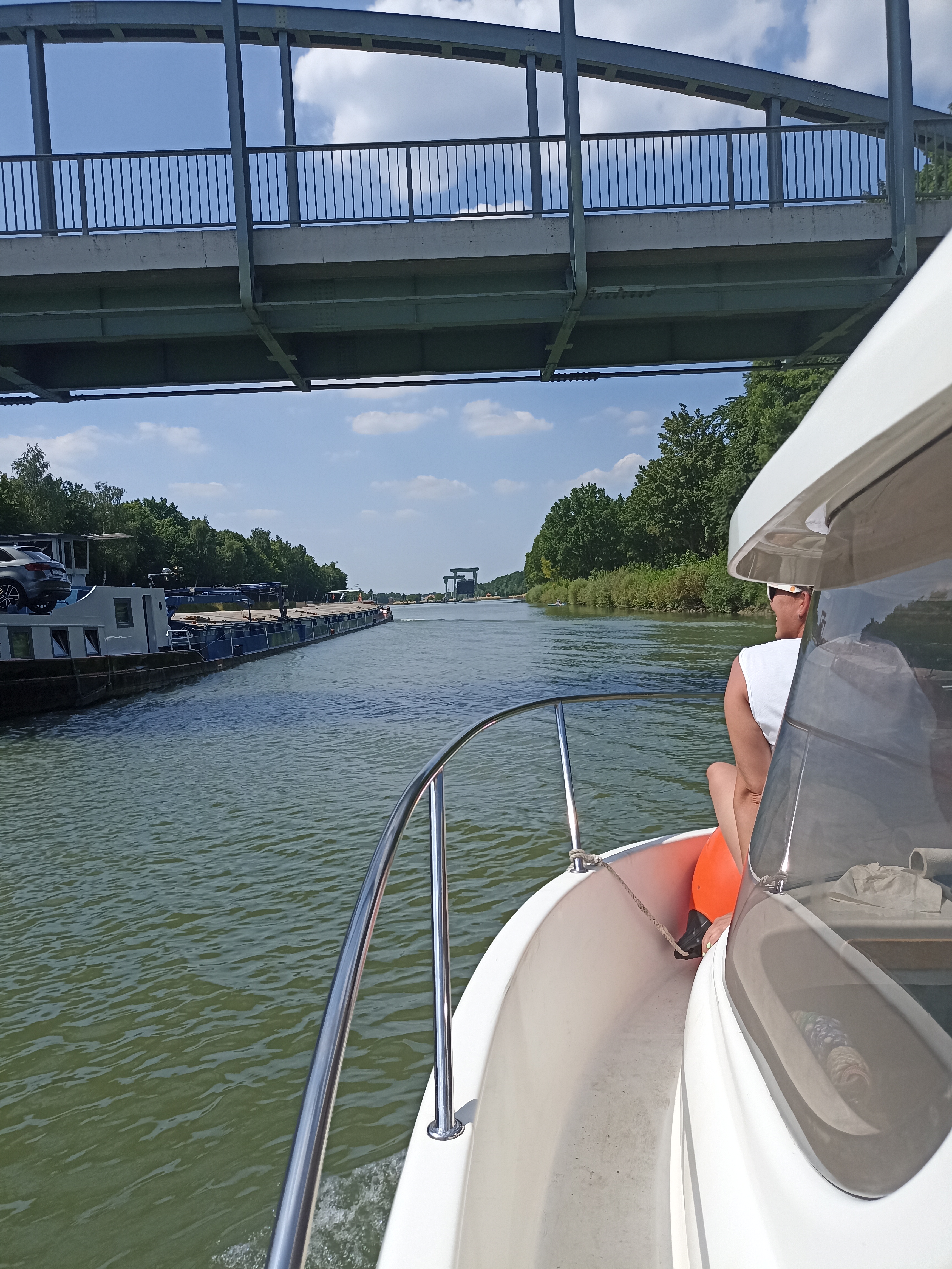 Auf dem Kanal wird ein Sportboot von einem Binnenschiff überholt, im Bug sitzt eine weibliche Person, am Horizont ist eine Schleuse zu sehen
