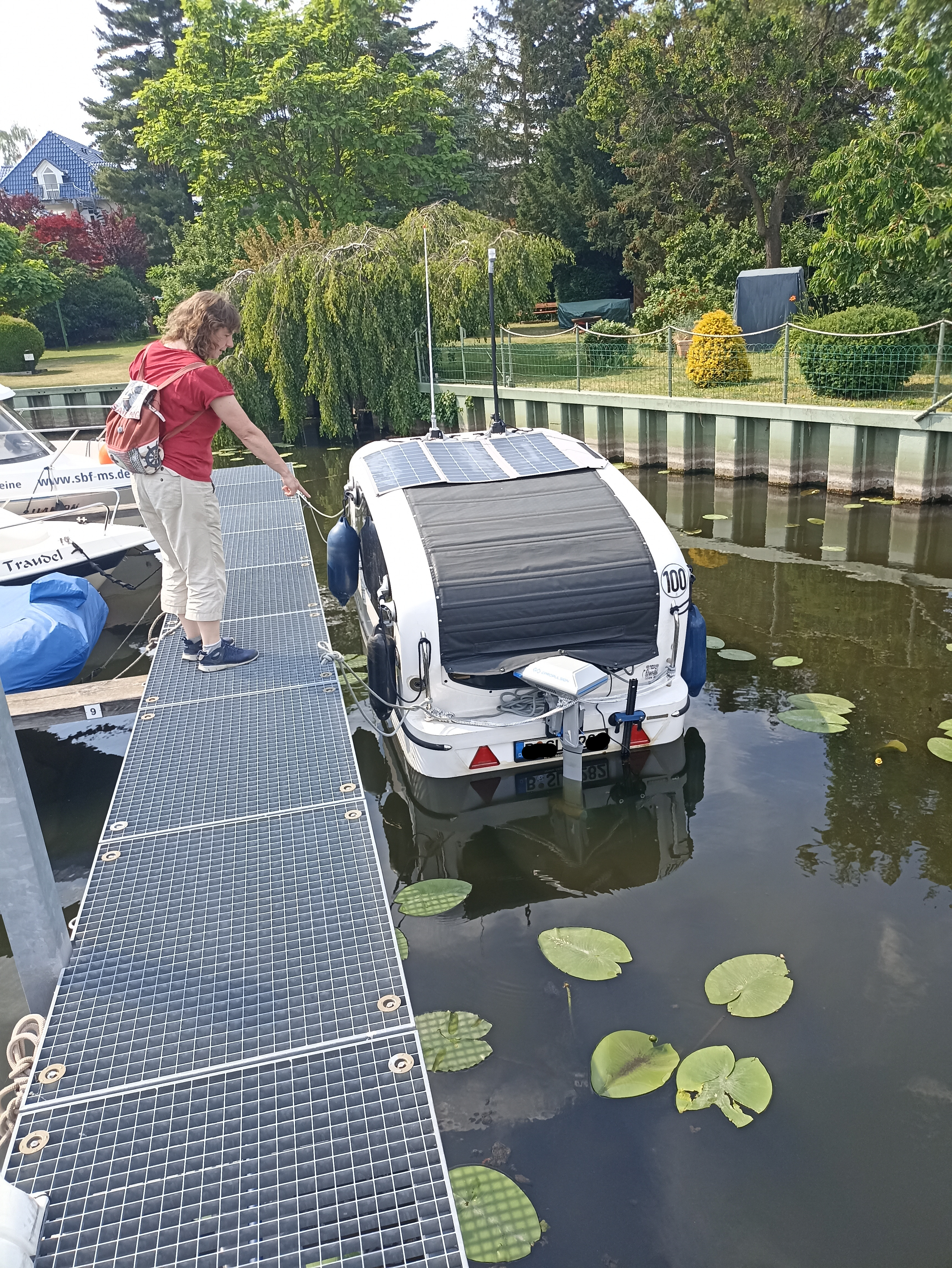 Ein Boot-Wohnwagen von der Rückseite im Wasser, am Steg, daneben eine weibliche Person die auf das Boot mit der Hand deutet.