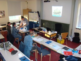In einem Schulungsraum sitzen drei Personen an Tischen, drauf liegen Blätter und Bücher, eine Person steht vor den Tischen und erklärt mit den Händen